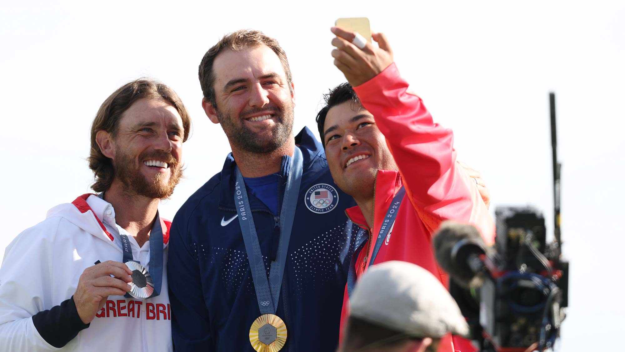 Gold medalist, Scottie Scheffler of Team United States (C), Silver medalist, Tommy Fleetwood of Team Great Britain (L) and Bronze medalist, Hideki Matsuyama of Team Japan (R) pose for a selfie on the podium during the Men's Individual Stroke Play medal ceremony following Day Four of the Men's Individual Stroke Play on day nine of the Olympic Games Paris 2024 at Le Golf National 